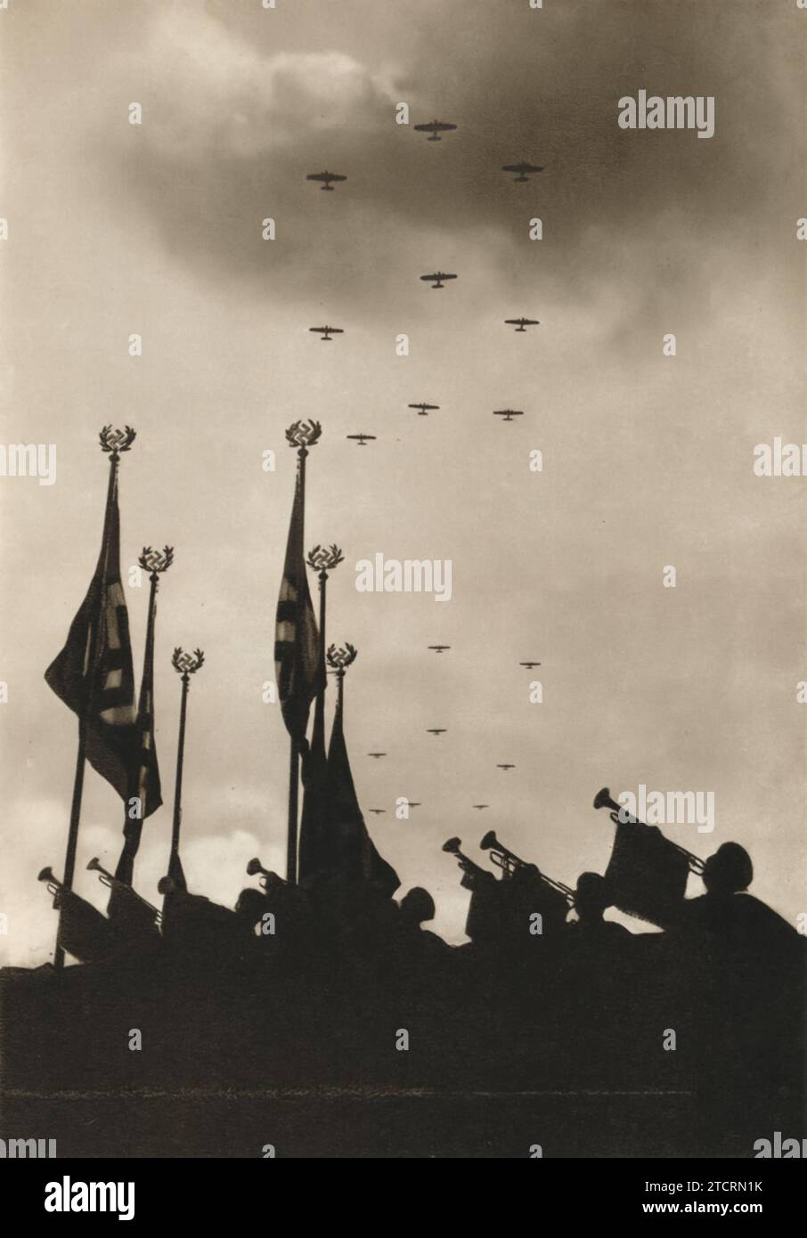 Dieses Bild zeigt eine deutsche Parade mit Fahnen und Trompeten im Vordergrund, die Nationalstolz und militärische Macht symbolisieren. Im Hintergrund sieht man die Luftwaffe, die als Luftwaffe bekannt ist, in die Luft aufsteigen und der Parade eine dynamische Luftdimension verleihen. Das begleitende Zitat "Luftfahrt ist eine Notwendigkeit" unterstreicht die entscheidende Rolle der Luftmacht, die das deutsche Militär während des Zweiten Weltkriegs anerkannt hat Stockfoto