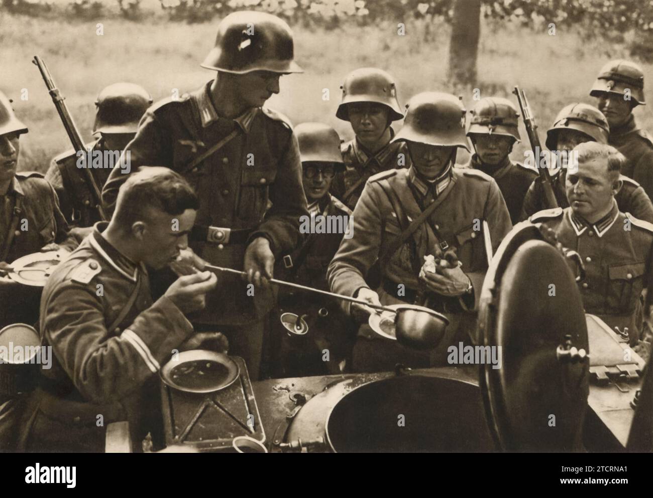 Selbst der Anführer findet das Essen ausgezeichnet. Diese Szene zeigt einen hochrangigen deutschen Offizier, der die gleichen militärischen Rationen wie seine Truppen schätzt. Es ist ein Moment, in dem sich die Ranggrenzen über ein gemeinsames Erlebnis verwischen und die Kameradschaft und den universellen Reiz einer guten Mahlzeit inmitten des Militärlebens hervorheben. Stockfoto