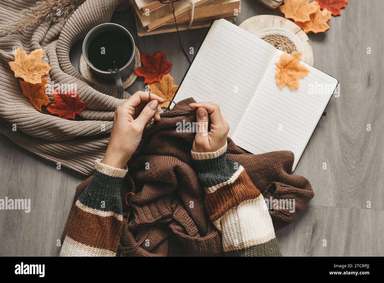 Weibliche Hände stricken Pullover auf dem Boden mit Herbstblättern und einer Tasse Kaffee Stockfoto