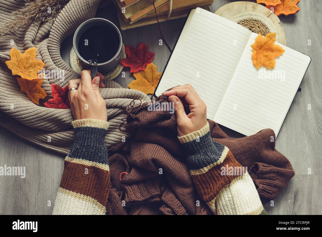 Weibliche Hände stricken Pullover auf dem Boden mit Herbstblättern und einer Tasse Kaffee Stockfoto