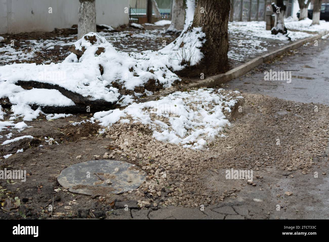 Unerledigte Straßenreparaturen. Kanalluke und gebrochener Asphalt, bedeckt mit zerquetschtem Stein. Schmutz und Schnee auf einer kaputten Straße in einem Wohngebiet. Flach Stockfoto