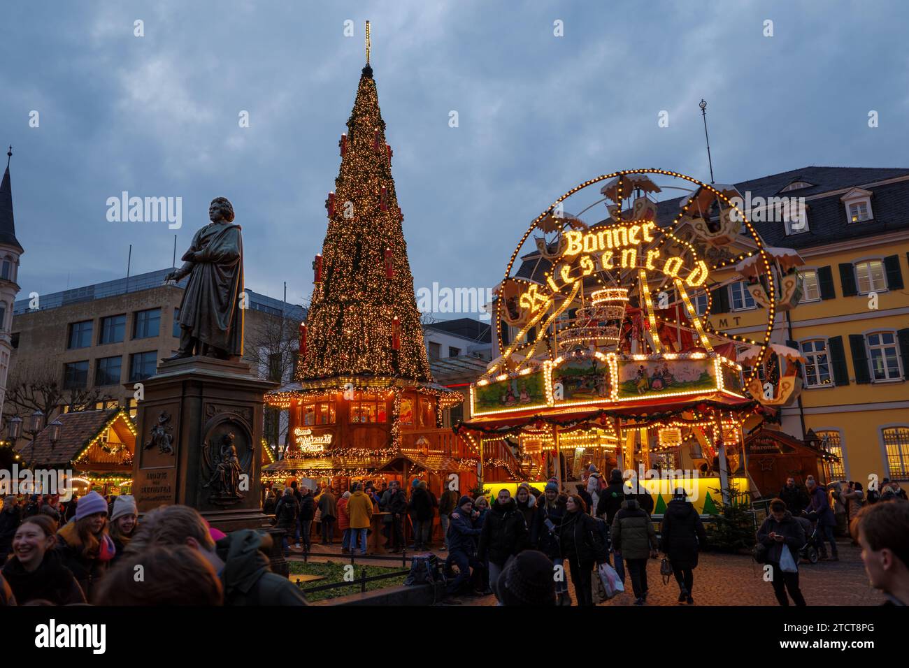 Bonn, Deutschland - 6. Dezember 2023: Blick in die Dämmerung auf einen belebten Weihnachtsmarkt mit festlichen Lichtern, einem Karussell und einem großen geschmückten Weihnachtsbaum dahinter Stockfoto