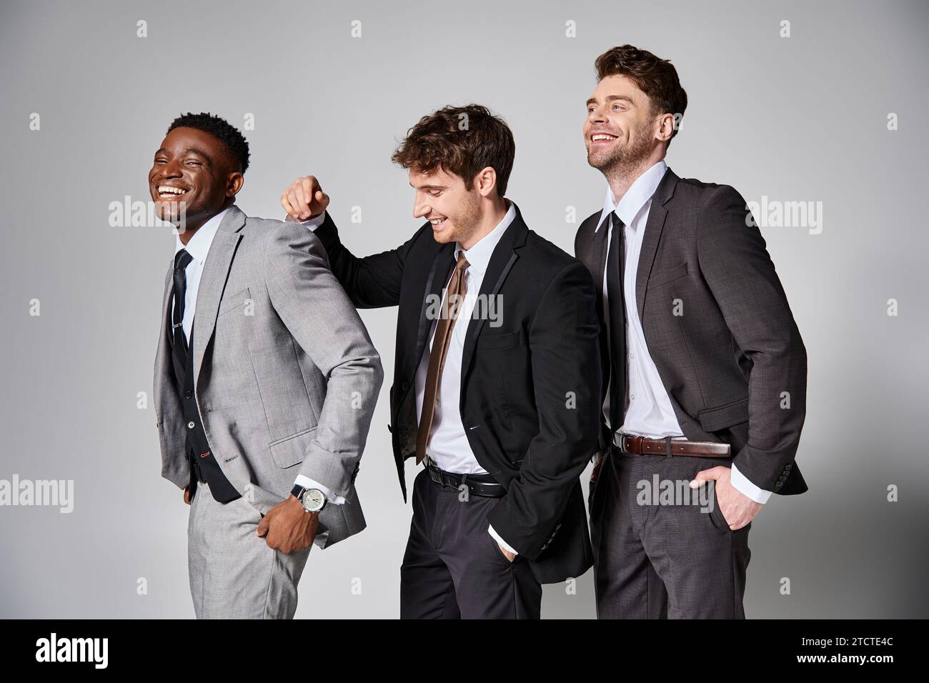 Attraktive, glückliche multikulturelle männliche Models in eleganten Anzügen, die aufrichtig vor grauem Hintergrund lächeln Stockfoto
