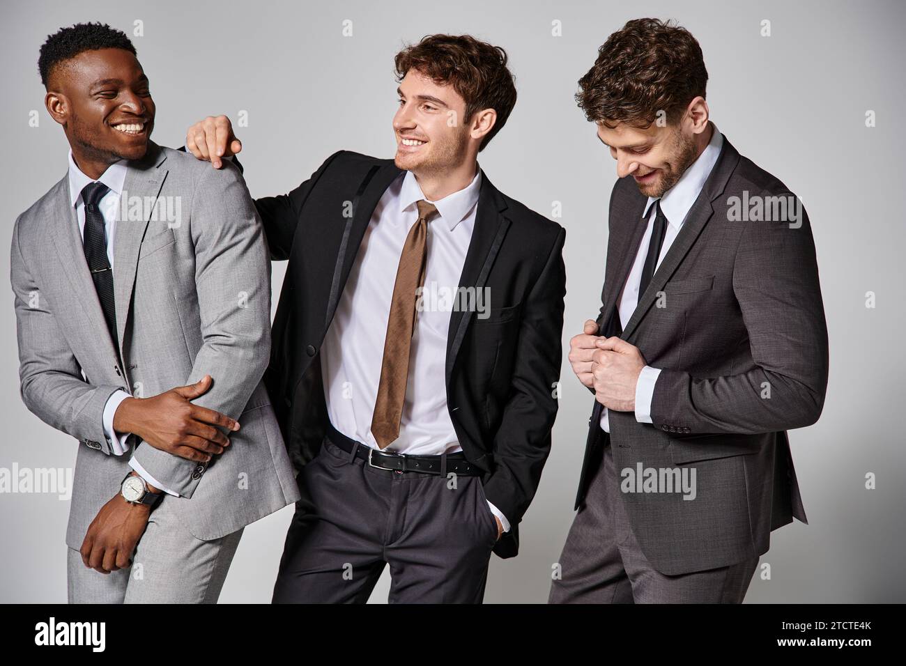 Gut aussehende, fröhliche, interrassische männliche Models in schicken Anzügen, die aufrichtig vor grauem Hintergrund lächeln Stockfoto