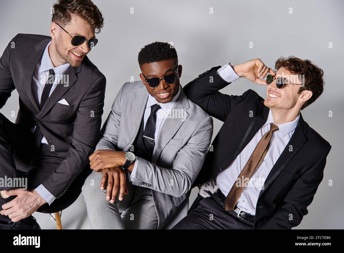 Fröhliche attraktive multikulturelle Männer in Business-Anzügen sitzen auf Stühlen und lächeln fröhlich Stockfoto
