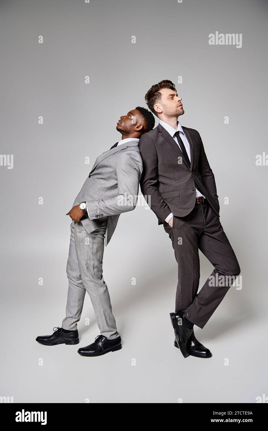 Zwei attraktive, stilvolle, multikulturelle männliche Models in Business-Kleidung, die vor grauem Hintergrund posieren Stockfoto
