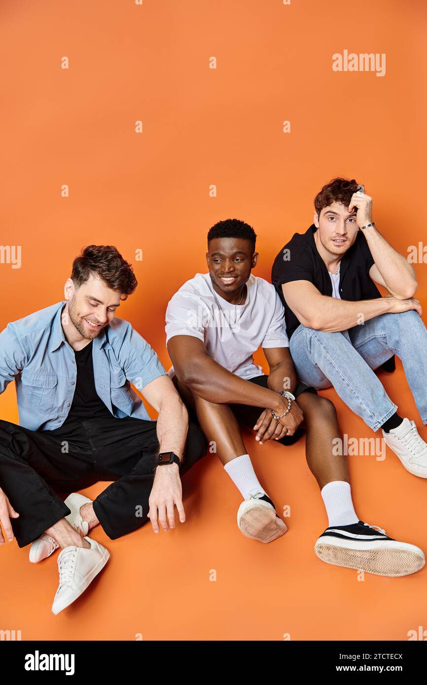 Drei fröhliche multirassische Männer in urbaner Freizeitkleidung lächelnd und sitzend auf dem Boden, Modekonzept Stockfoto