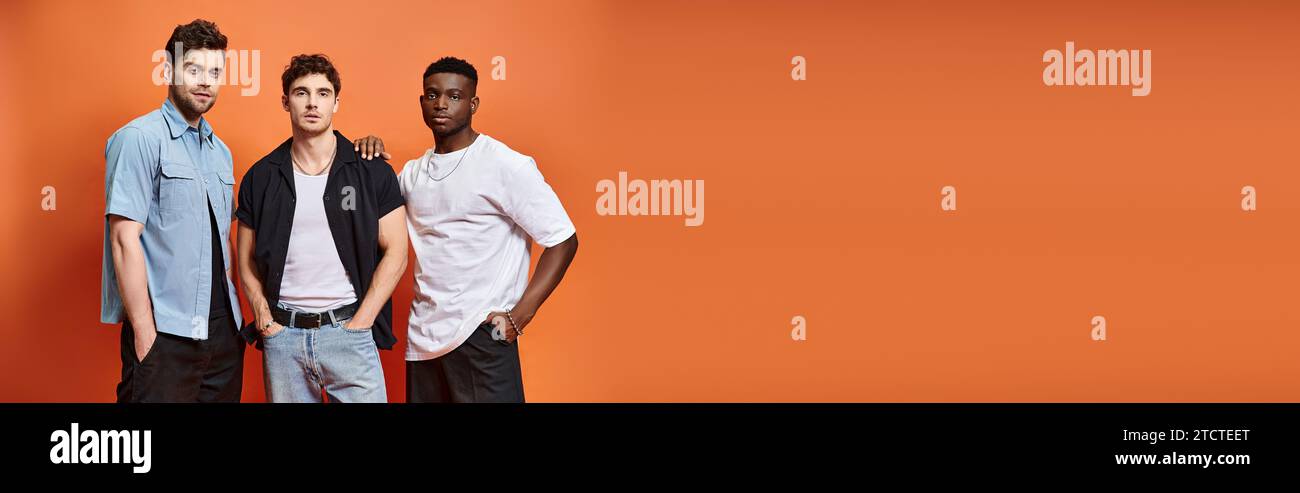 Hübsche, vielseitige Männer in trendigen urbanen Kleidern, die vor orangefarbenem Hintergrund posieren, Modekonzept, Banner Stockfoto