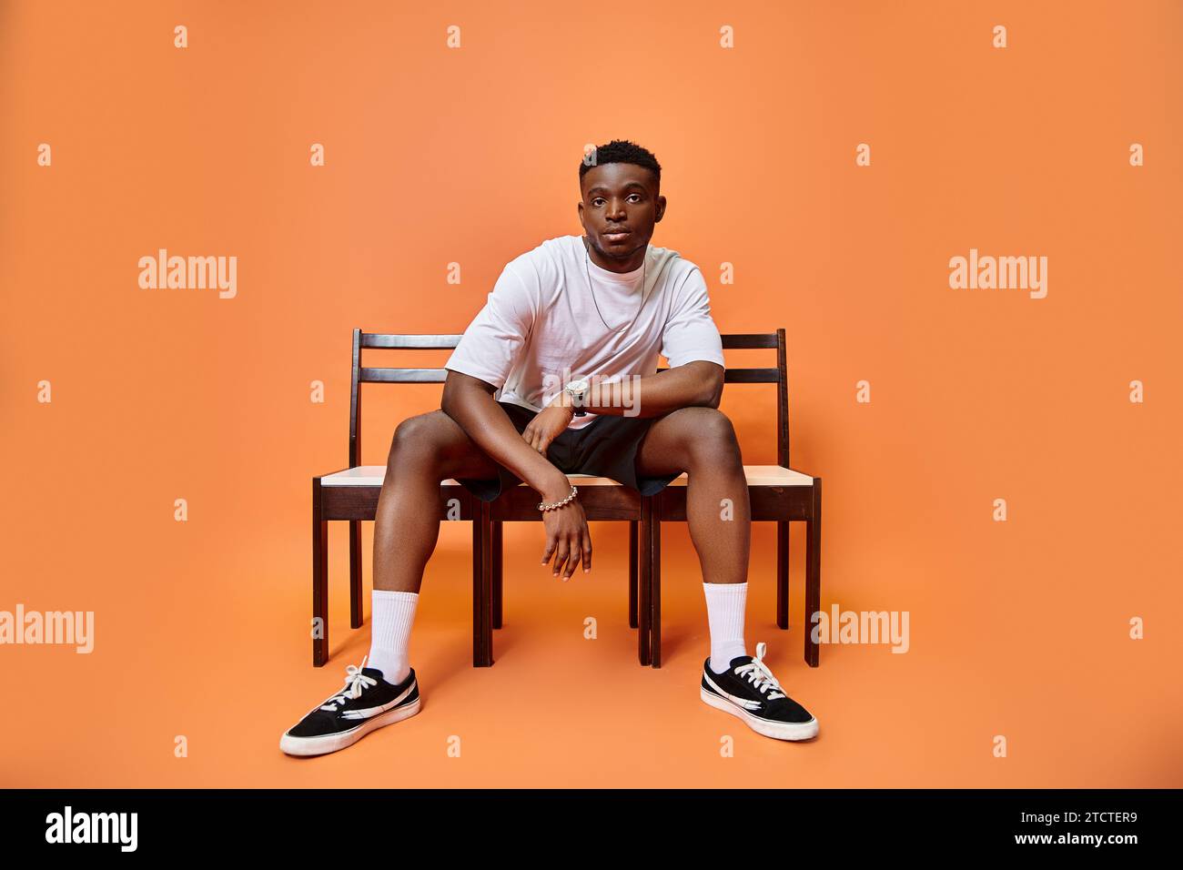 Ein ansprechender junger afroamerikaner in urbanem Outfit, der auf einer Bank vor orangefarbenem Hintergrund sitzt, Mode Stockfoto