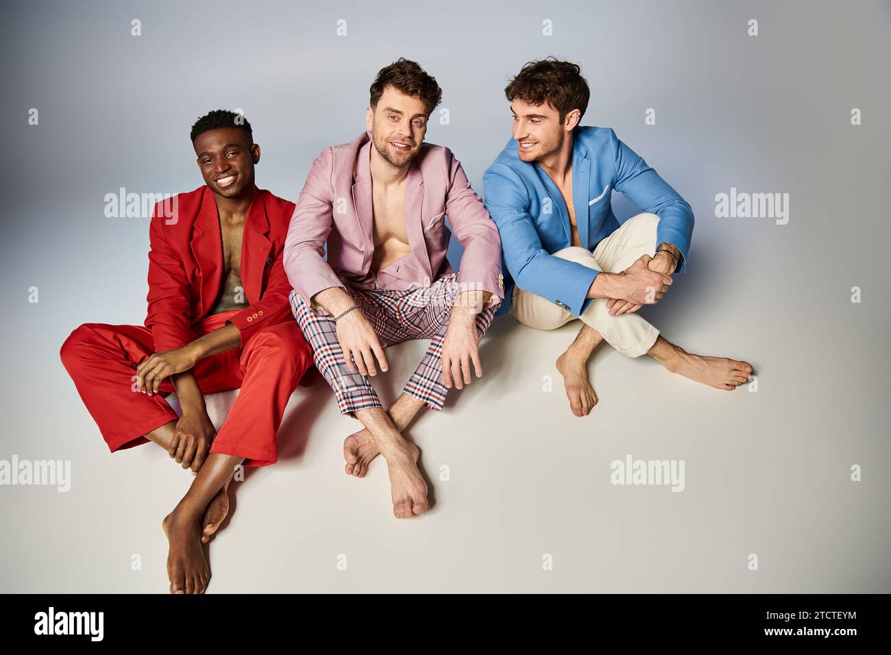 Fröhliche interrassische Freunde in farbenfrohen Anzügen, die mit überkreuzten Beinen auf dem Boden sitzen, Mode Stockfoto