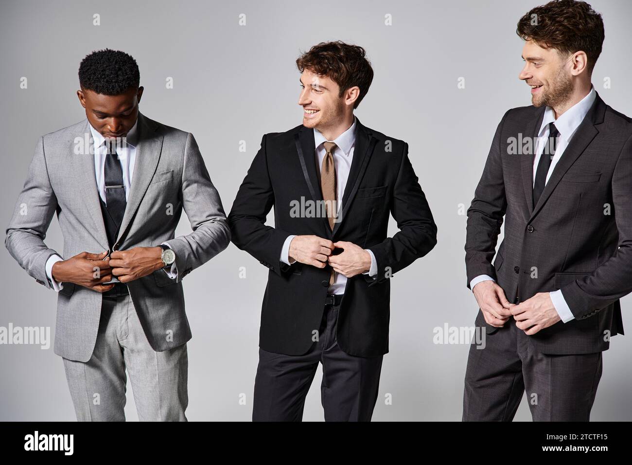 Ansprechende, multikulturelle männliche Models in eleganten Anzügen, die aufrichtig vor grauem Hintergrund lächeln Stockfoto