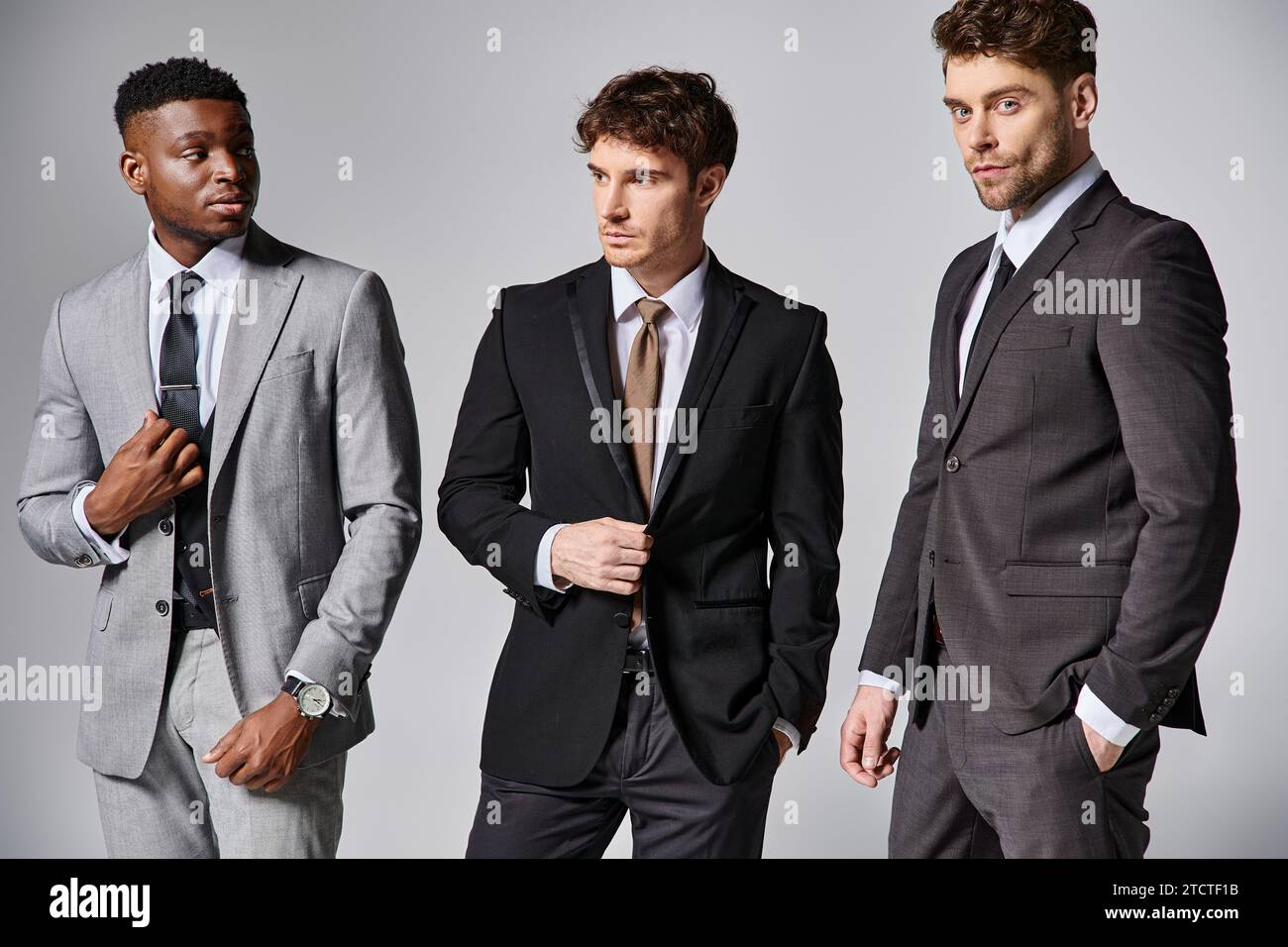 Hübsche, stilvolle, multikulturelle männliche Models in Business-eleganten Anzügen, die vor grauem Hintergrund posieren Stockfoto