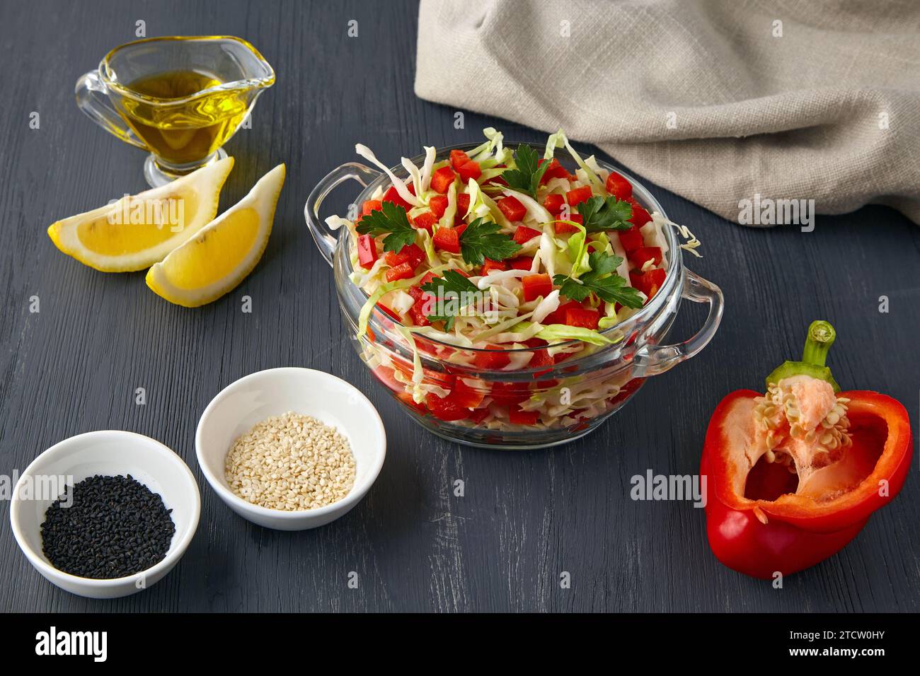 Frischer Gemüsesalat aus Kohl und süßen Paprika in einer Glasschale auf schwarzem hölzernem Hintergrund, umgeben von Zutaten Stockfoto