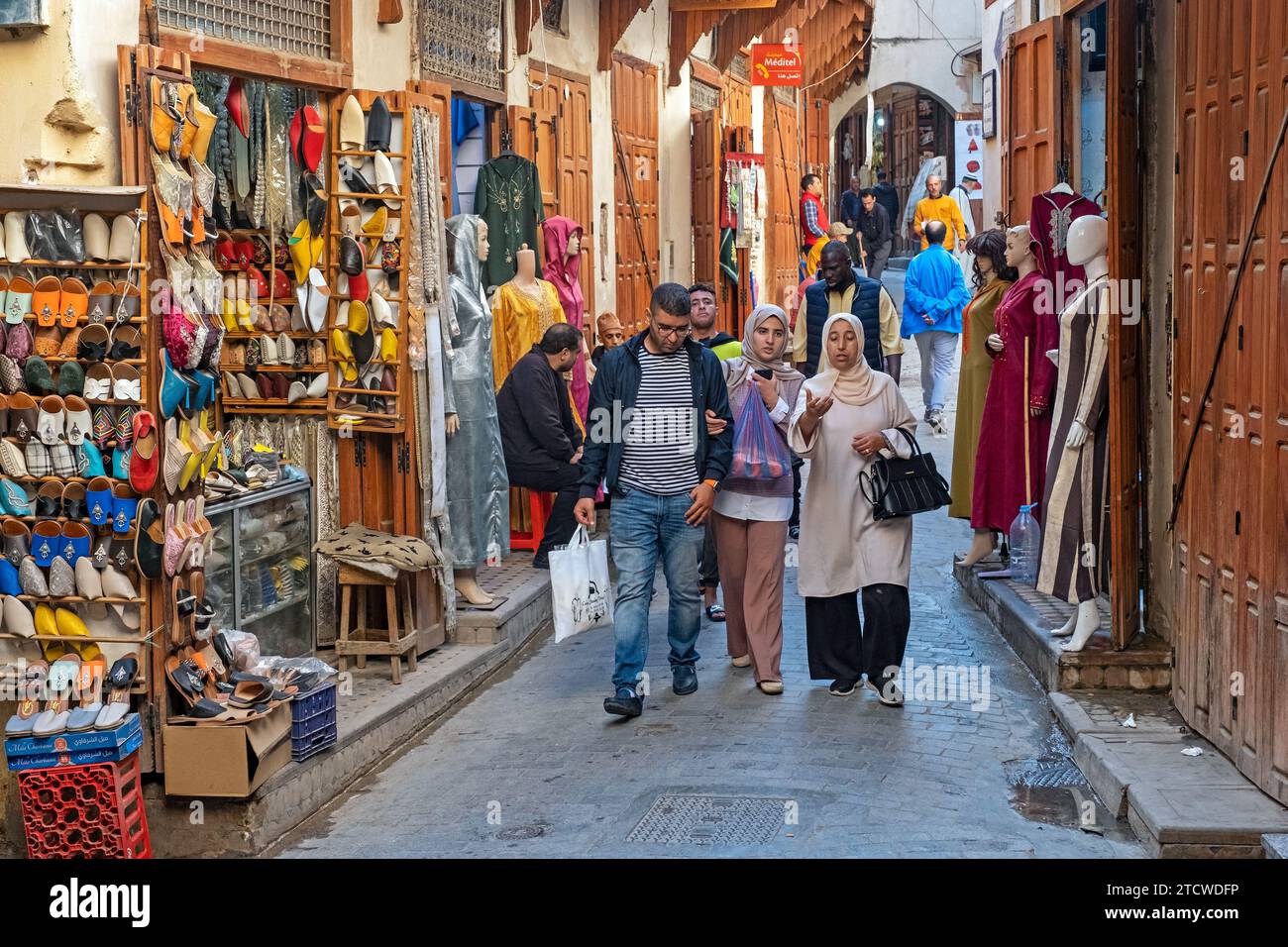 Marokkaner laufen in der Einkaufsstraße vorbei an Schuhladen und Kleiderladen in Medina in der Stadt Fes / Fès, Fès-Meknes, Marokko Stockfoto