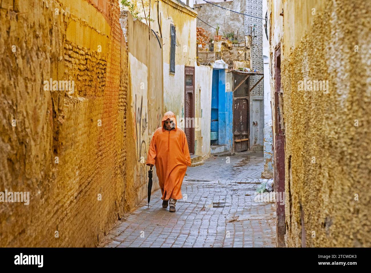 Muslimischer Mann trägt orange Djellaba / Jillaba an einem regnerischen Tag in der Gasse der gelben Medina der Stadt Meknes, Fez-Meknes, Marokko Stockfoto