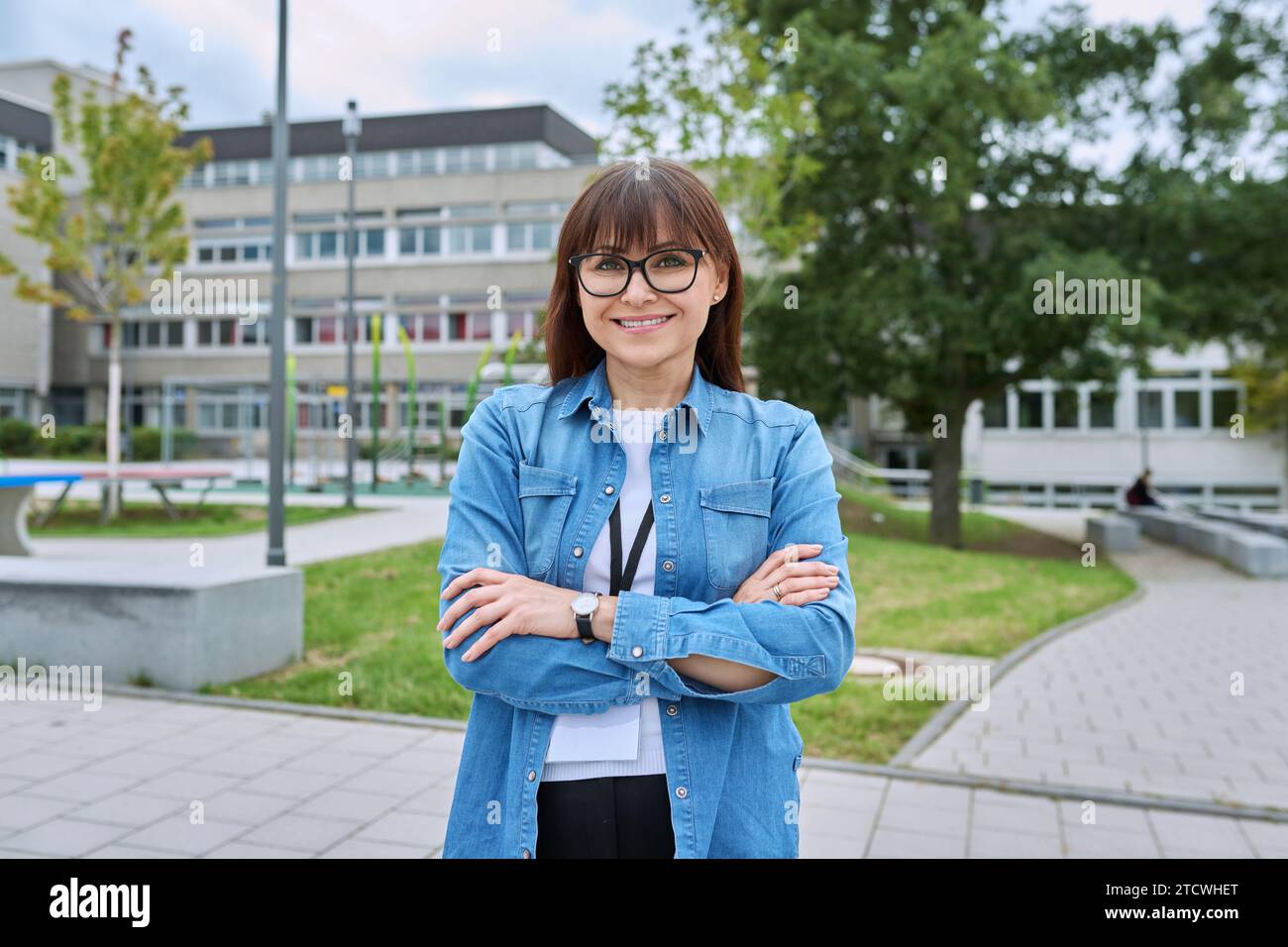 Selbstbewusste Lehrerin mittleren Alters mit gekreuzten Armen in der Nähe des Schulgebäudes Stockfoto