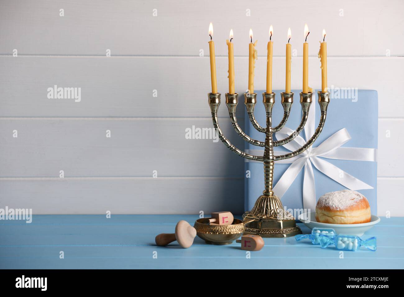 Hanukkah-Feier. Menora mit brennenden Kerzen, Dreidels, Donut und Geschenkbox auf hellblauem Holztisch, Platz für Text Stockfoto