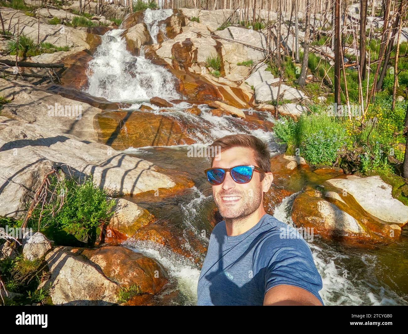 Fröhlicher Wanderer macht ein Selfie mit einem beeindruckenden Wasserfall im regenerierenden Wald des Rocky Mountain National Park. Stockfoto