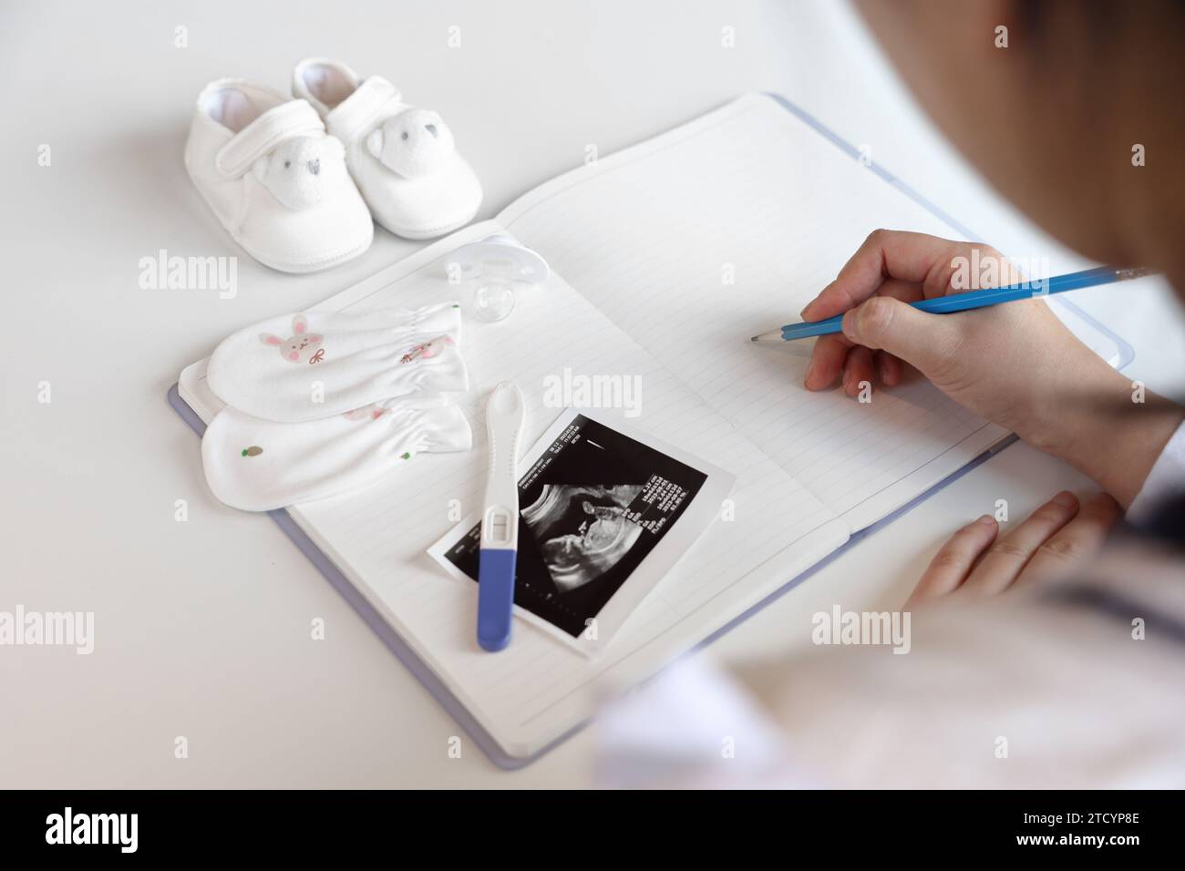 Eine schwangere Frau macht Notizen in ein leeres Notizbuch, Babyartikel und Ultraschallbilder werden platziert Stockfoto