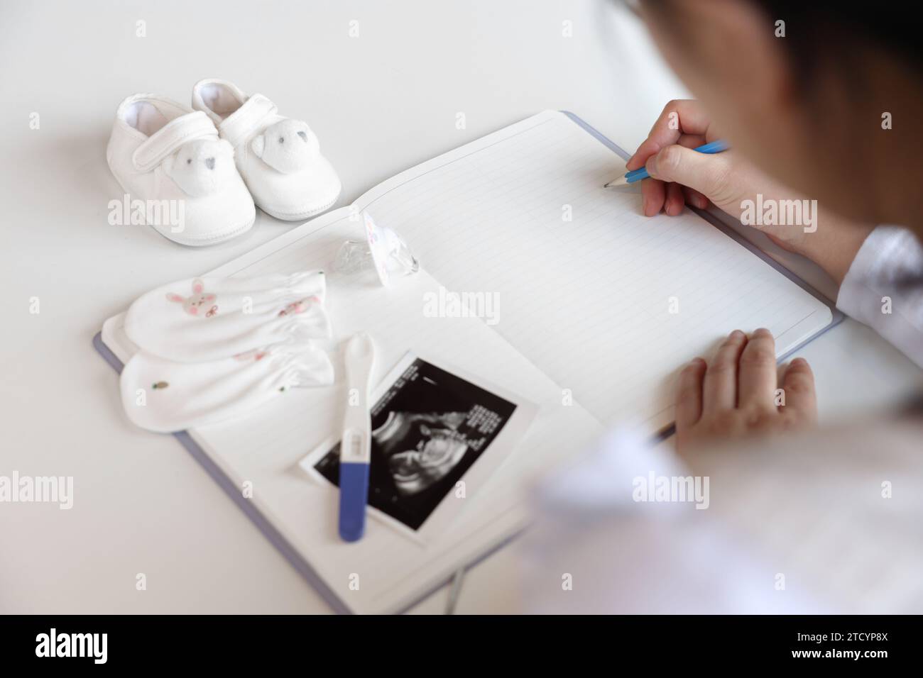Eine schwangere Frau macht Notizen in ein leeres Notizbuch, Babyartikel und Ultraschallbilder werden platziert Stockfoto