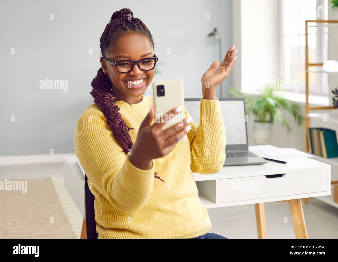 Glückliches lächelndes afroamerikanisches schönes Mädchen schaut auf ihr Handy, stehend im Wohnzimmer vor dem Hintergrund des Schreibtisches mit Laptop. Stockfoto