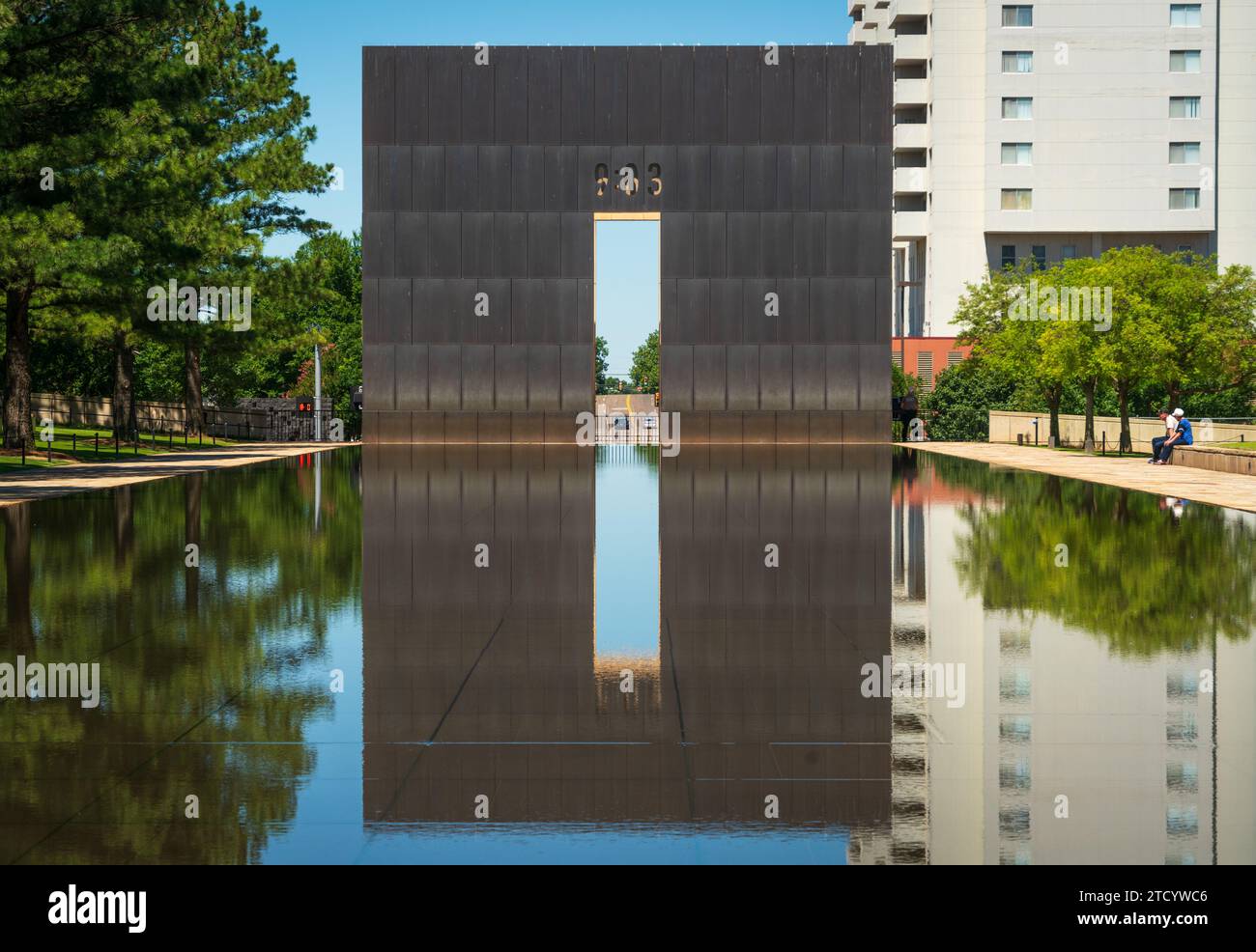 Das dunkle Tor mit seinem hellen Mittelschlitz spiegelt sich im noch spiegelnden Teich neben grünen Bäumen am Oklahoma City National Memorial, Oklahoma, wider. Stockfoto