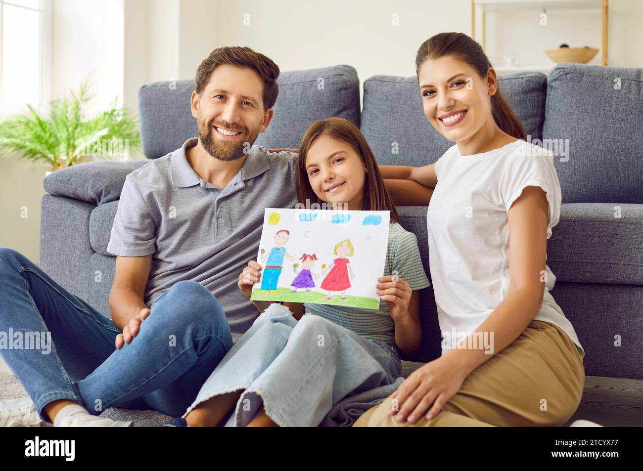 Lächelndes Mädchen, das auf dem Boden sitzt und die Eltern zu Hause zeigen, wie eine glückliche Familie gezeichnet ist. Stockfoto