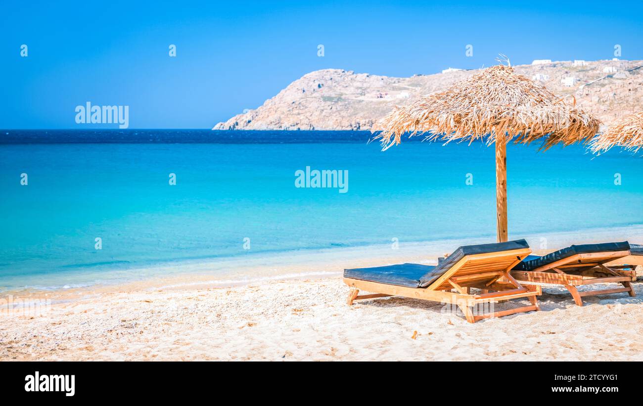 Mykonos Strand im Sommer mit Sonnenschirm und luxuriösen Liegestühlen, blaues Meer mit einem Berg am Elia Strand Mikonos Griechenland Strand im Sommer in Europa Stockfoto