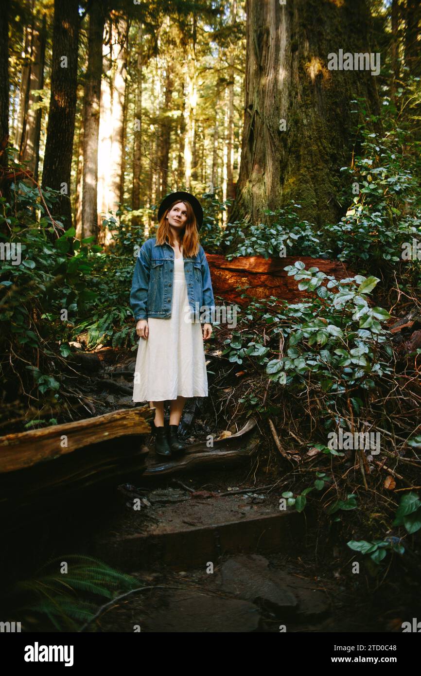 Eine junge Frau in lässigem Outfit und Hut steht auf einem Waldweg umgeben von hohen Bäumen auf Avatar Grove, Vancouver Island. Stockfoto