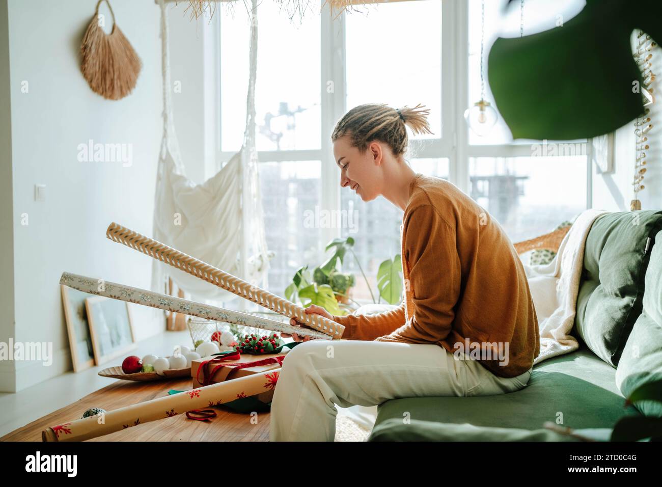 Seitenansicht einer jungen Frau in legeren Kleidern, die auf dem Sofa sitzt und zu Hause die Verpackung für Weihnachtsgeschenke in der Hand hält Stockfoto