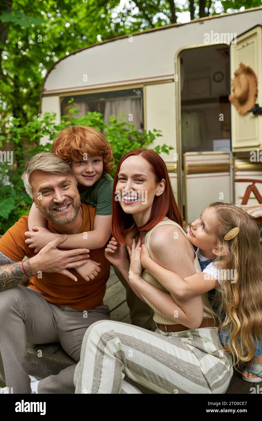 Aufgeregte Kinder, die lachende Eltern umarmen, die draußen im Wohnwagen sitzen, emotionale Verbindung Stockfoto