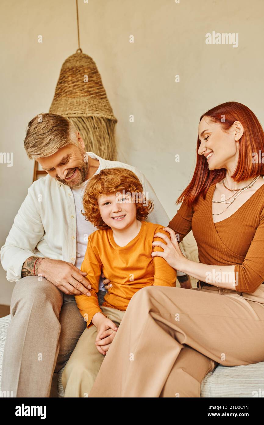 Lächelnder Ehemann und Ehefrau umarmen rothaariges Kind, während sie im Schlafzimmer sitzen, emotionale Verbindung Stockfoto
