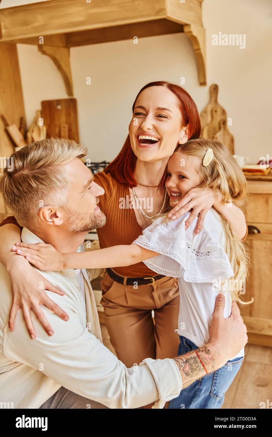 Fröhliche Eltern mit entzückender Tochter, die sich in der gemütlichen Küche zuhause umarmen, verbindende Momente Stockfoto