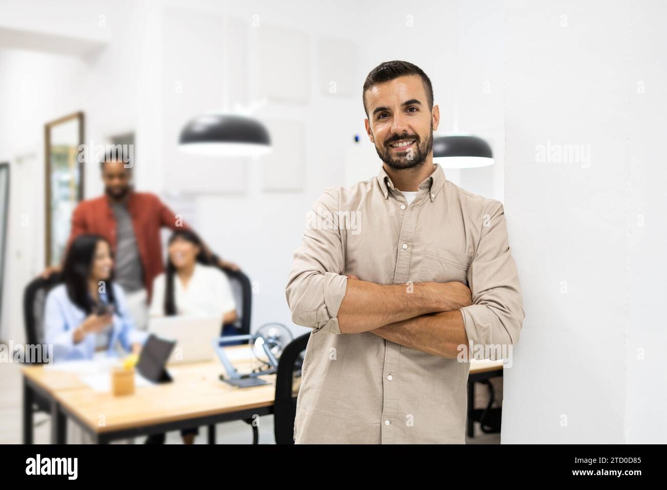 Porträt eines lächelnden, selbstbewussten jungen kaukasischen Geschäftsmannes in Casuals mit überkreuzten Armen, der an die Wand gelehnt ist im kreativen Büro Stockfoto