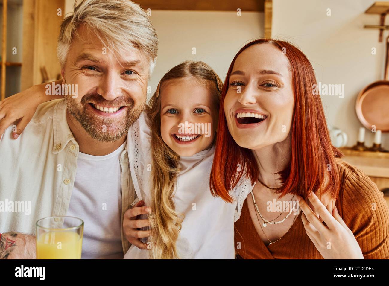 Aufgeregte Eltern mit süßer Tochter, die in der Küche zu Hause in die Kamera schaut, familiäre Interaktion Stockfoto