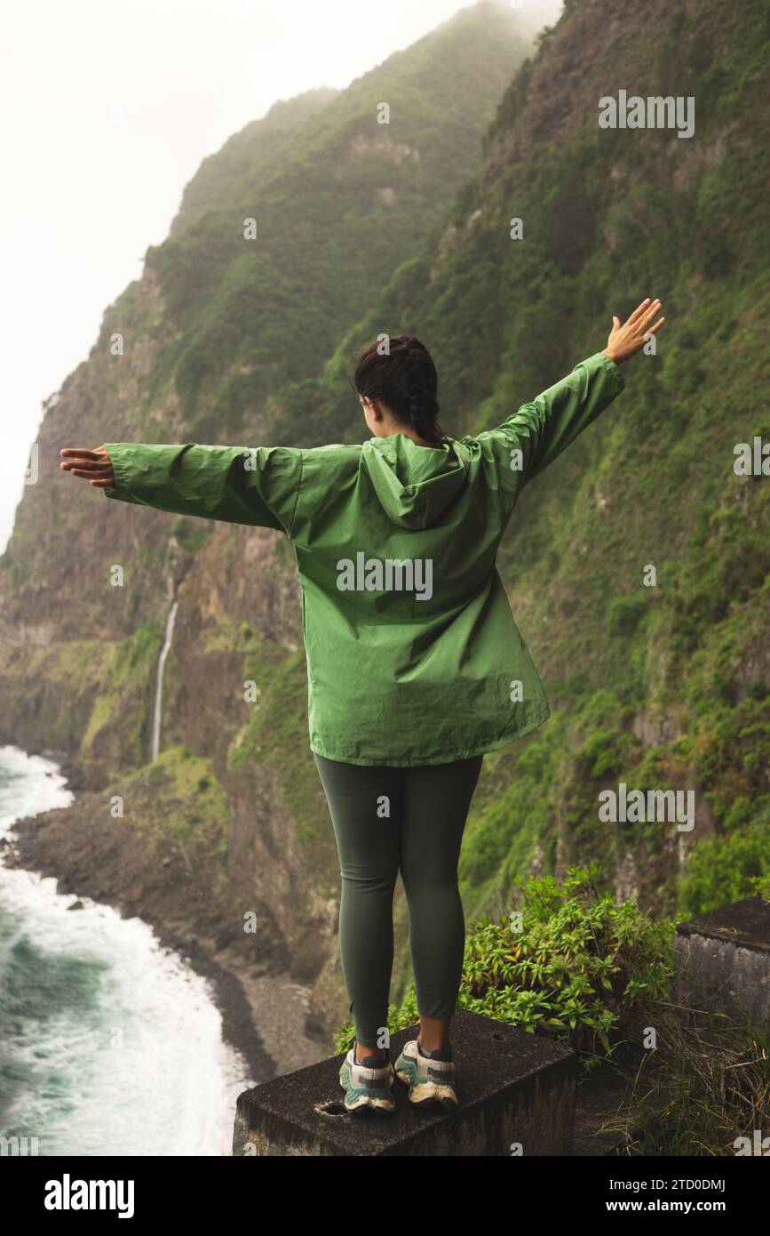 Rückansicht einer unbedenklichen, nicht erkennbaren jungen Frau in grüner Regenjacke, die mit ausgerückten Armen auf Betonbarriere gegen Berge steht Stockfoto