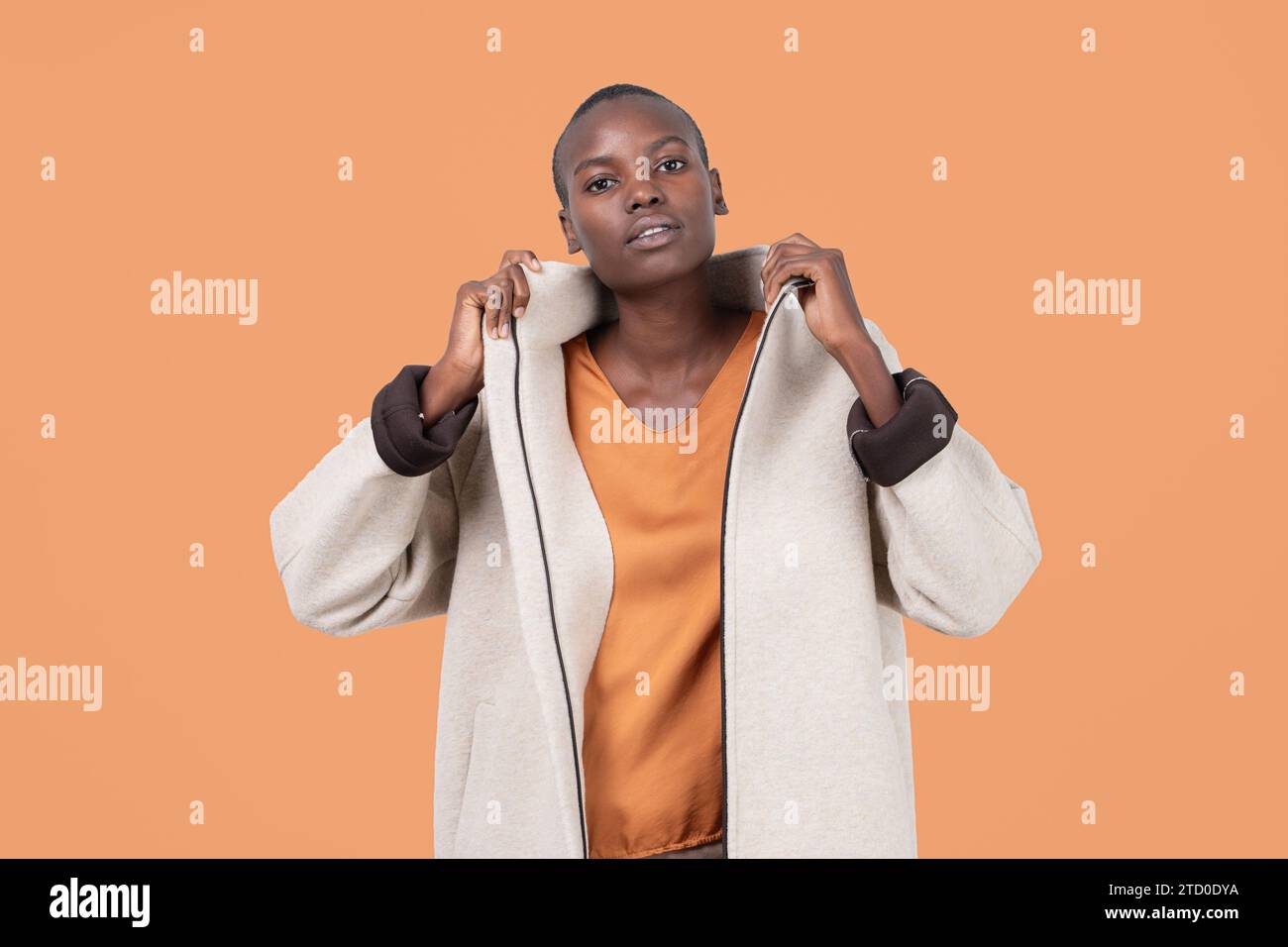 Porträt einer jungen, mutigen afroamerikanischen Frau im langen Reißverschluss-Mantel, die selbstbewusst in die Kamera blickt, die auf orangefarbenem Hintergrund steht Stockfoto