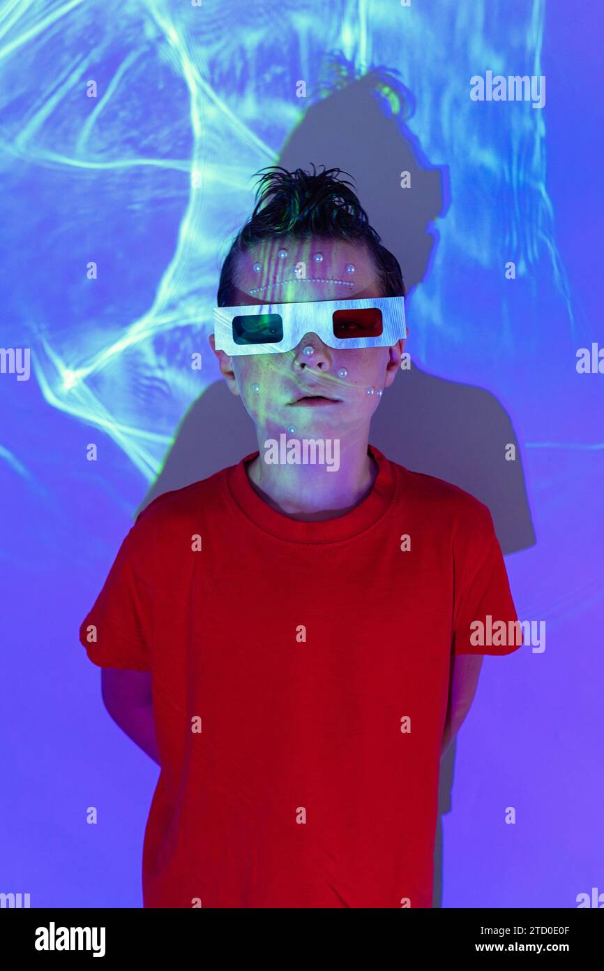 Porträt eines niedlichen Jungen in legeren Kleidern mit Perle im Gesicht und 3D-Brille, die vor der Wand mit leuchtenden blauen Neonlichtern steht, während sie auf den Camer blickt Stockfoto