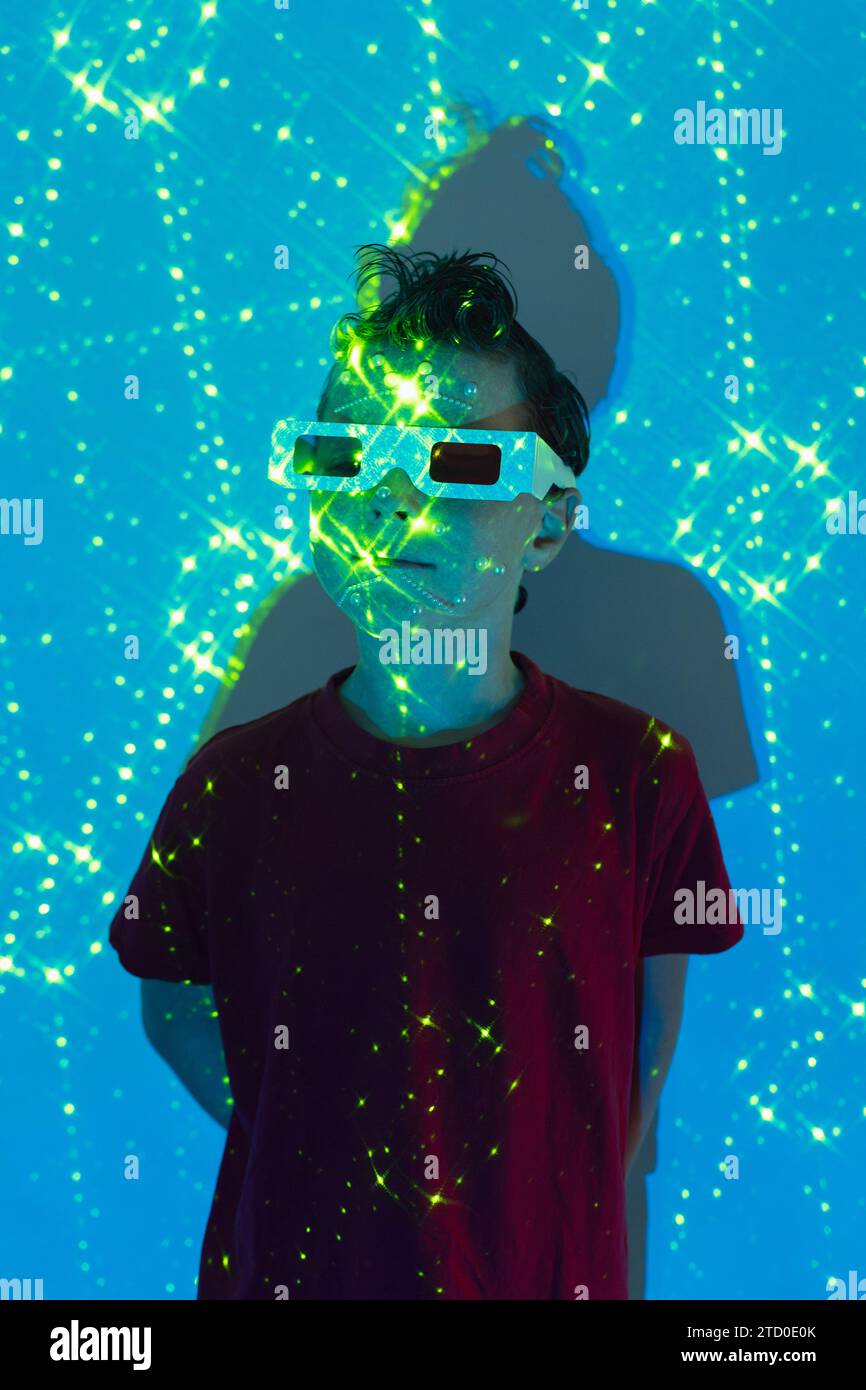 Porträt eines niedlichen Jungen in legeren Kleidern mit Perlen im Gesicht und 3D-Brille, die an der Wand mit leuchtenden blauen und grünen Neonlichtern steht Stockfoto