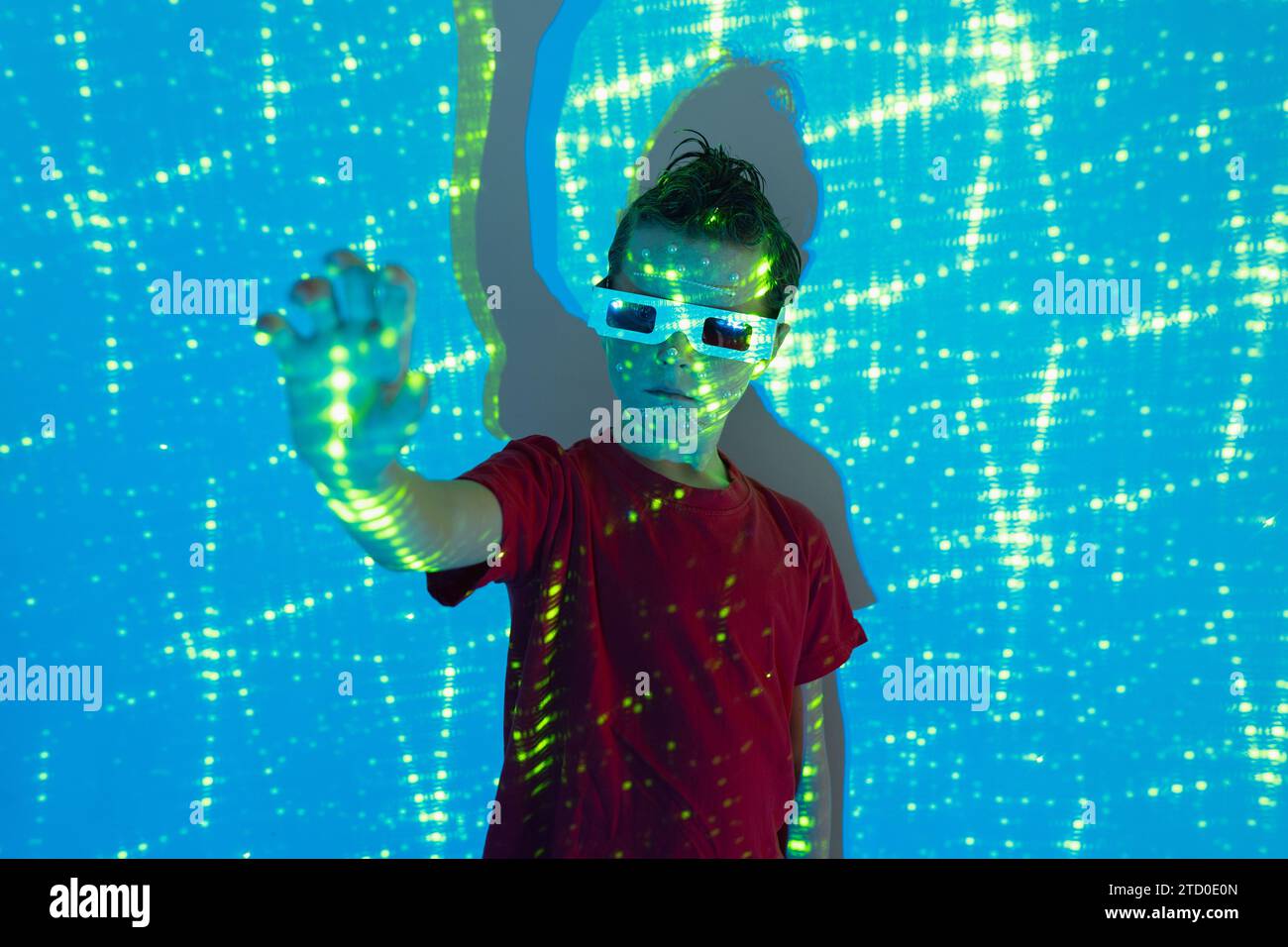 Porträt eines niedlichen Jungen in legeren Kleidern mit Perlen im Gesicht und 3D-Brille, die mit der Hand an der Wand steht, mit beleuchteten blauen Neonlichtern, während l Stockfoto
