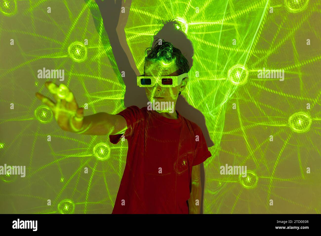 Porträt eines niedlichen Jungen in legeren Kleidern mit Perlen im Gesicht und 3D-Brille, die mit der Hand an der Wand steht und mit leuchtenden grünen Neonlichtern leuchtet Stockfoto