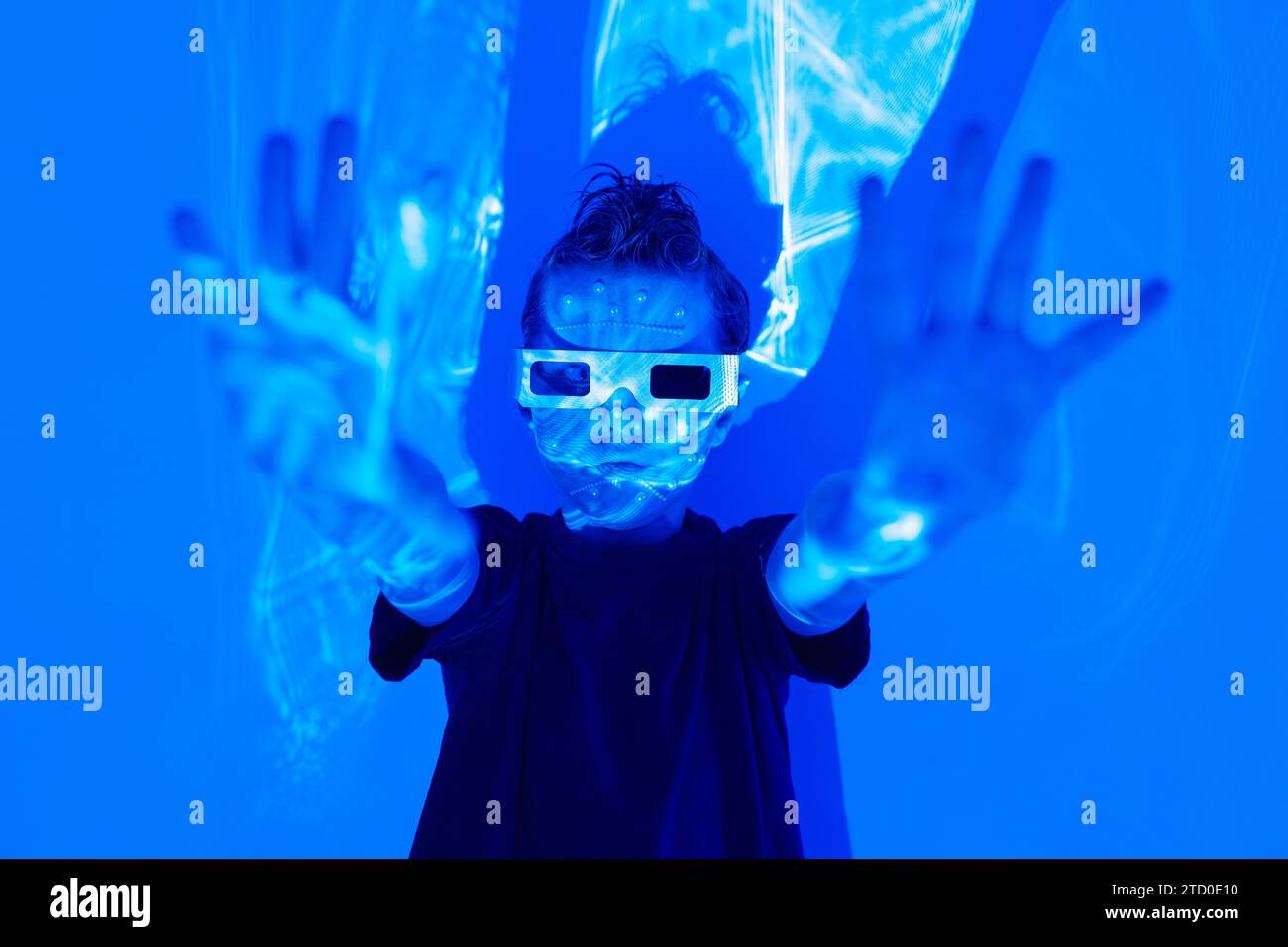Porträt eines selbstbewussten Jungen in legeren Kleidern mit Perlen im Gesicht und 3D-Brille, die mit den Händen nach außen im leuchtenden blauen Neonstudio vor der Wand steht Stockfoto