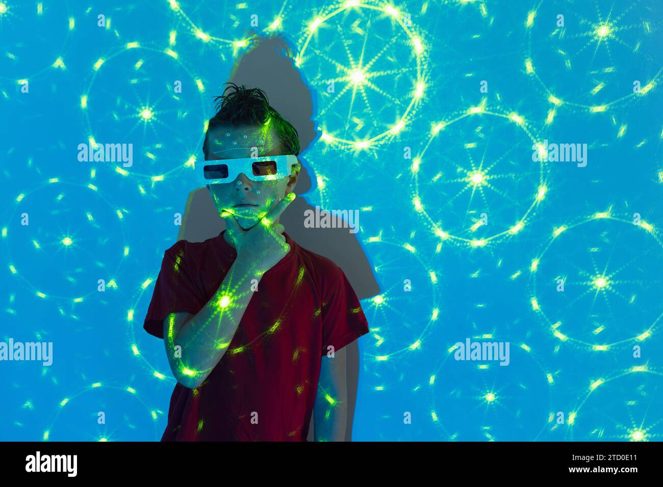 Porträt eines niedlichen Jungen in legeren Kleidern mit Perlen im Gesicht und 3D-Brille, die vor der Wand mit leuchtenden blauen Neonlichtern steht, während sie sich angeschaut hat Stockfoto