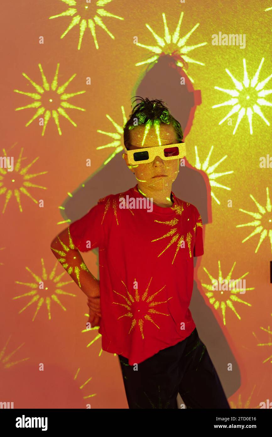 Niedlicher Junge in legeren Kleidern mit Perlen im Gesicht und 3D-Brille, die an der Wand mit leuchtenden gelben Neonlichtern steht, während er mit ha in die Kamera blickt Stockfoto