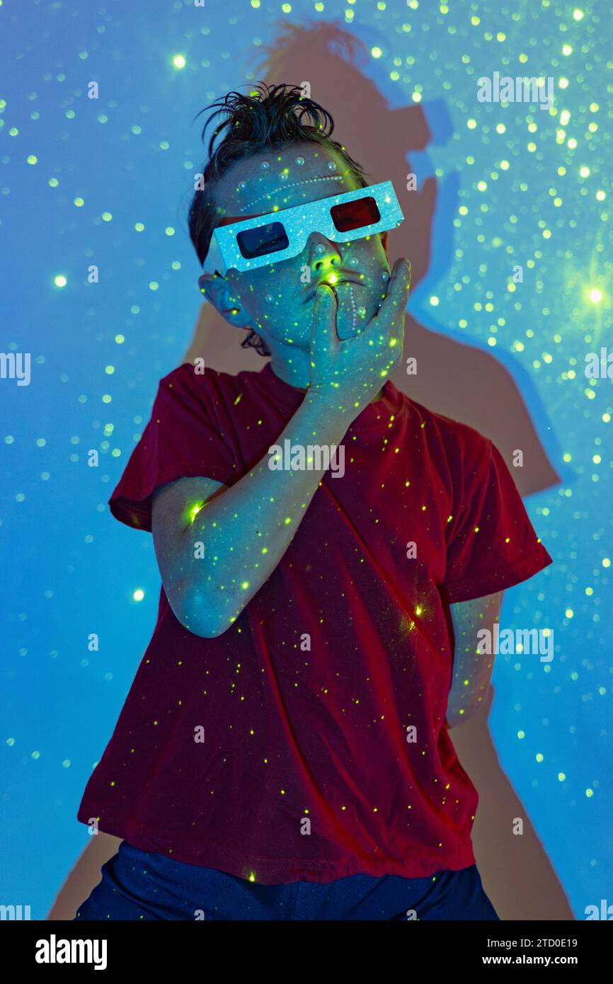 Selbstbewusster Junge in legeren Kleidern mit Perlen im Gesicht und 3D-Brille mit Hand am Kinn, die im Studio mit beleuchteten Neonlichtern an der Wand steht Stockfoto