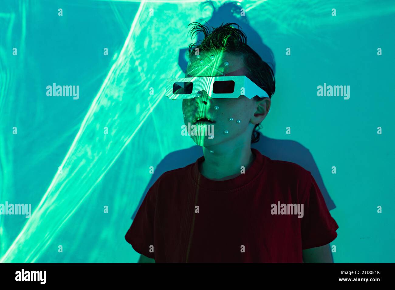 Porträt eines niedlichen Jungen in legeren Kleidern mit Perlen im Gesicht und 3D-Brille, die an der Wand mit leuchtenden blauen Neonlichtern steht und wegblickt Stockfoto