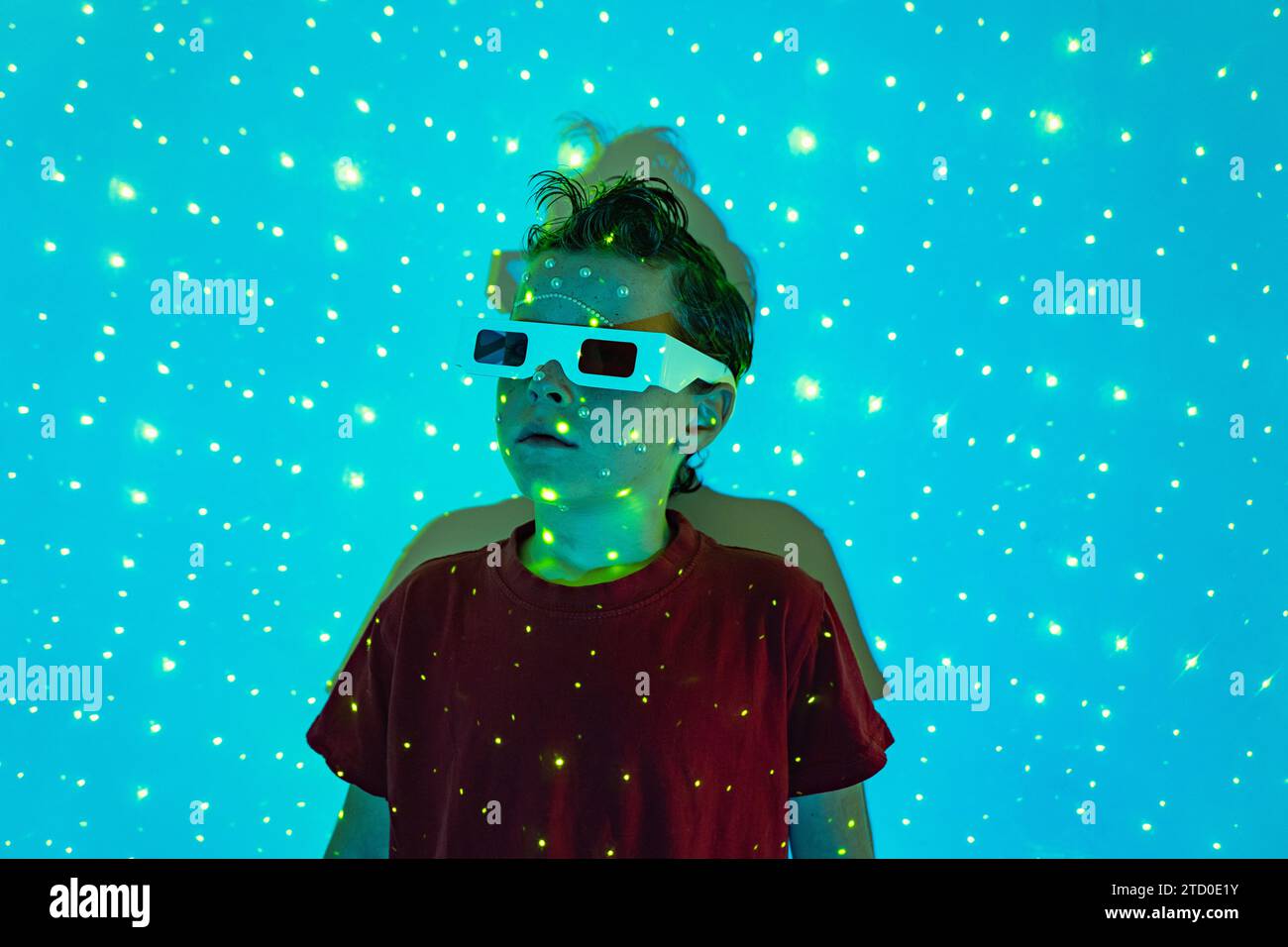 Porträt eines niedlichen Jungen in legeren Kleidern mit Perlen im Gesicht und 3D-Brille, die an der Wand mit leuchtenden blauen Neonlichtern steht und wegblickt Stockfoto