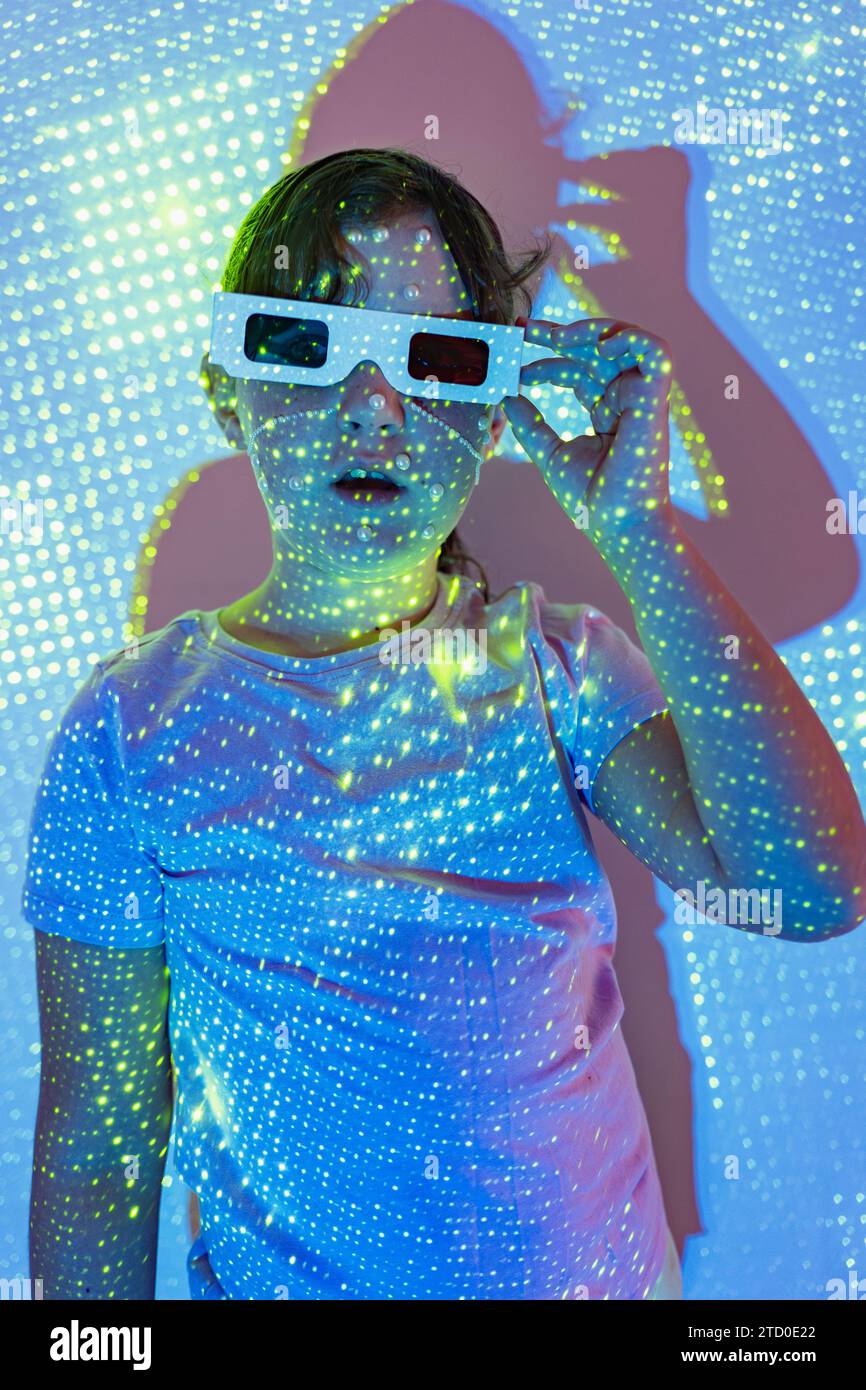 Porträt eines Mädchens in lässiger Kleidung und einer 3D-Brille mit Perlen im Gesicht, die in beleuchteten Neonlichtern im Studio steht, während sie mit geöffneter Kamera in die Kamera schaut Stockfoto