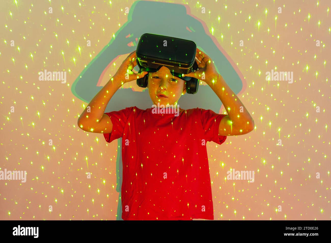 Porträt eines niedlichen Jungen in legeren Kleidern, der ein Virtual-Reality-Headset trägt und in die Kamera schaut, während er im Studio in gemusterten Neonlichtern steht Stockfoto