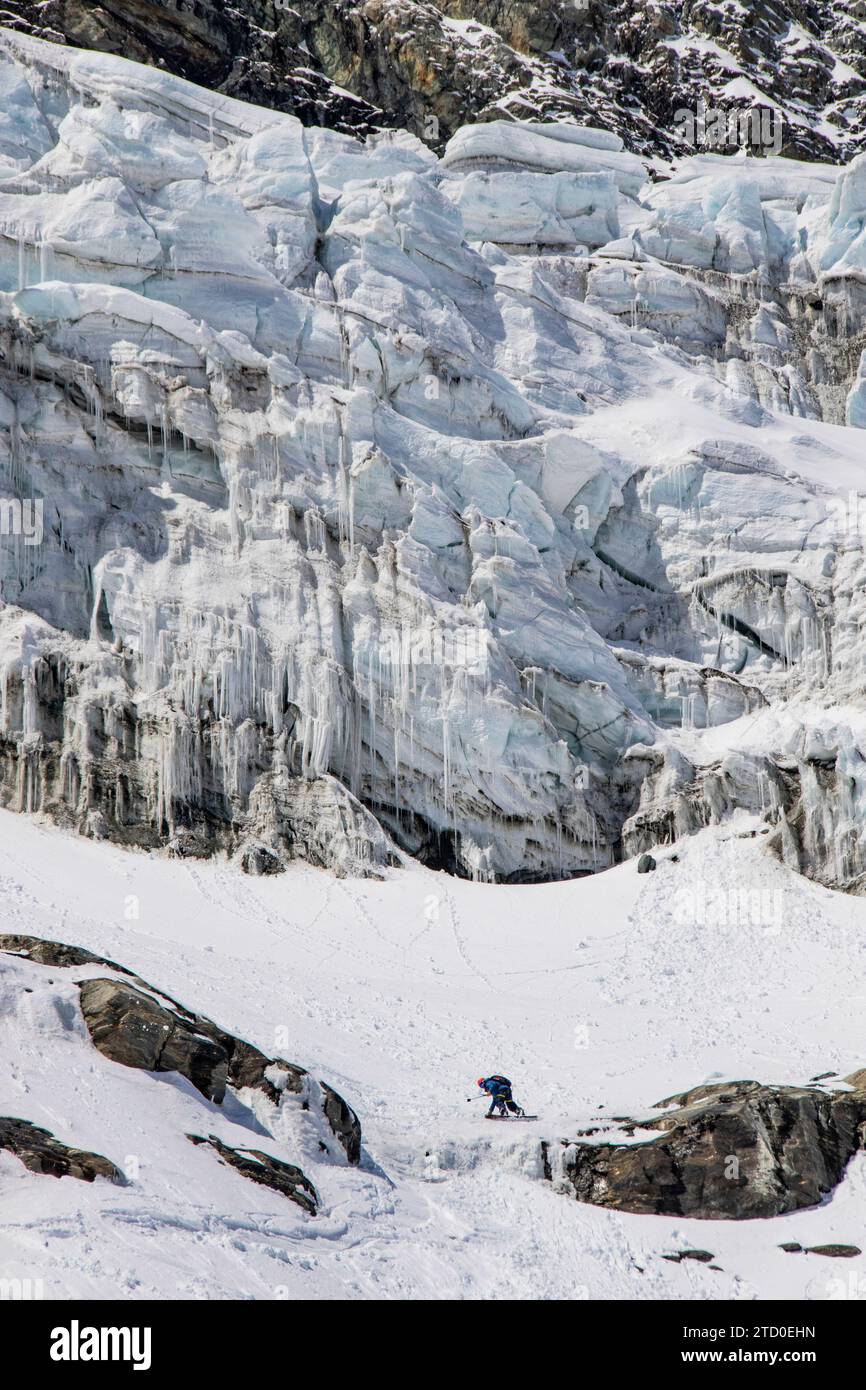Seitenansicht eines anonymen Skifahrers in lebendiger Kleidung im weitläufigen weißen Becken eines Gletschers, überschattet von den kolossalen Eismauern, die sich über ihnen erheben Stockfoto